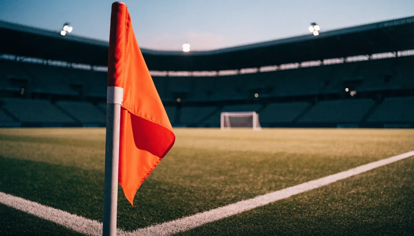 Eine Eckfahne am Rand eines Rasenplatzes, dahinter ein leeres Stadion unter Flutlicht aus der Ferne gesehen.