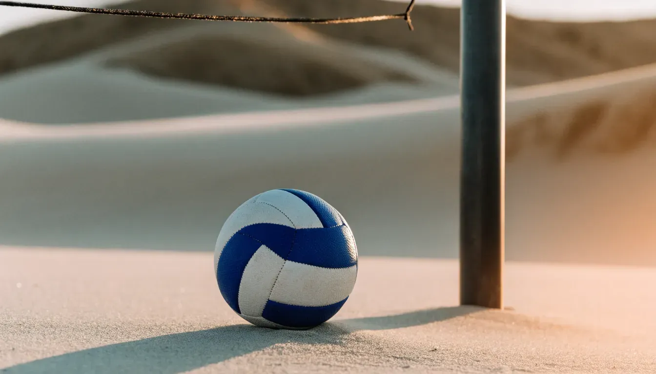 Ein Volleyball liegt auf Sand neben einem Netzpfosten, Strandlicht am späten Nachmittag, im Hintergrund unscharfe Dünen.
